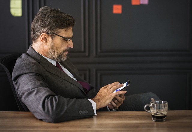 Muž v obleku sedí pri stole a pozerá do mobilného telefónu